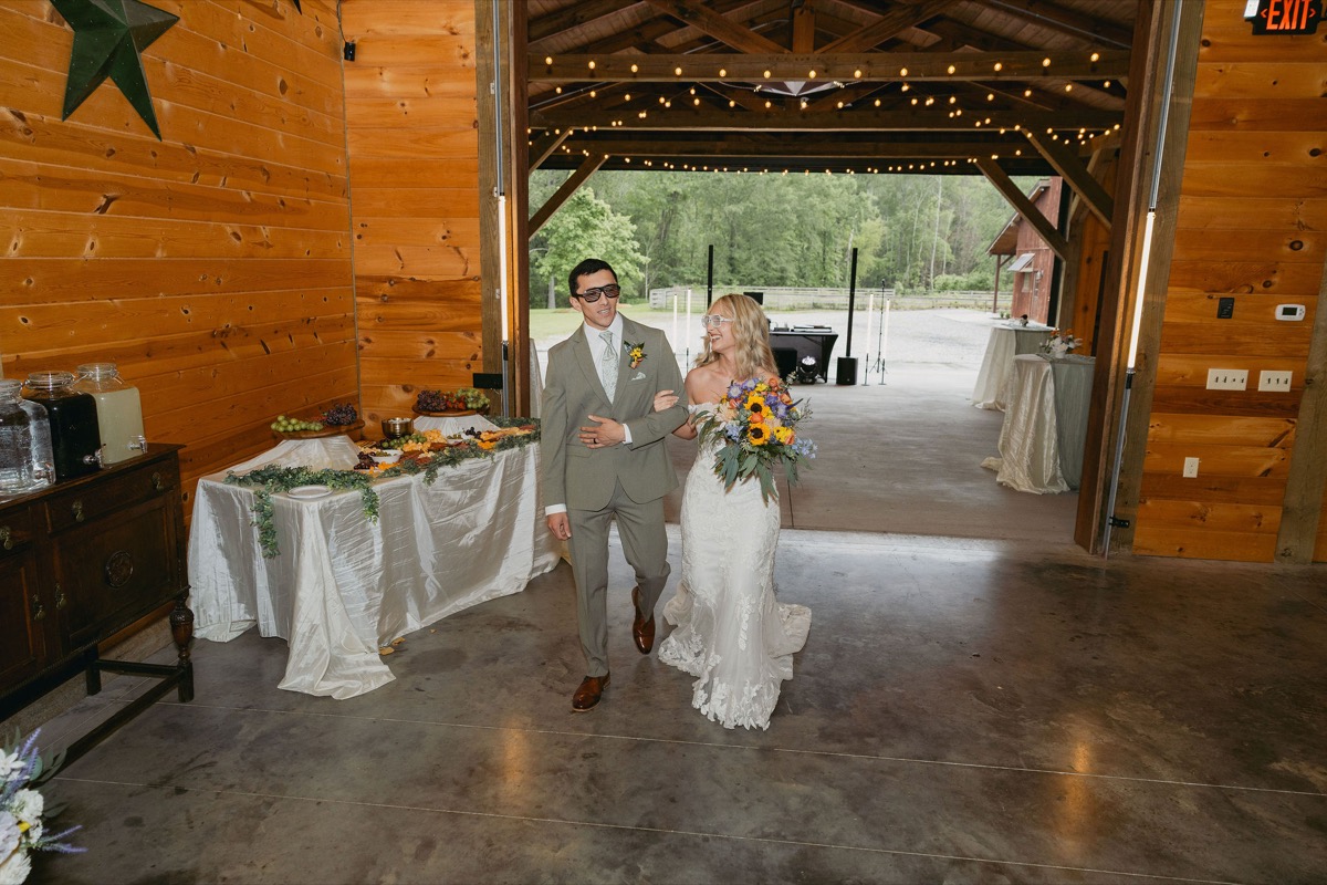 Brianna & Michael enter the string-lit barn in sunglasses, sunflower bouquet in hand, at Covey Creek.