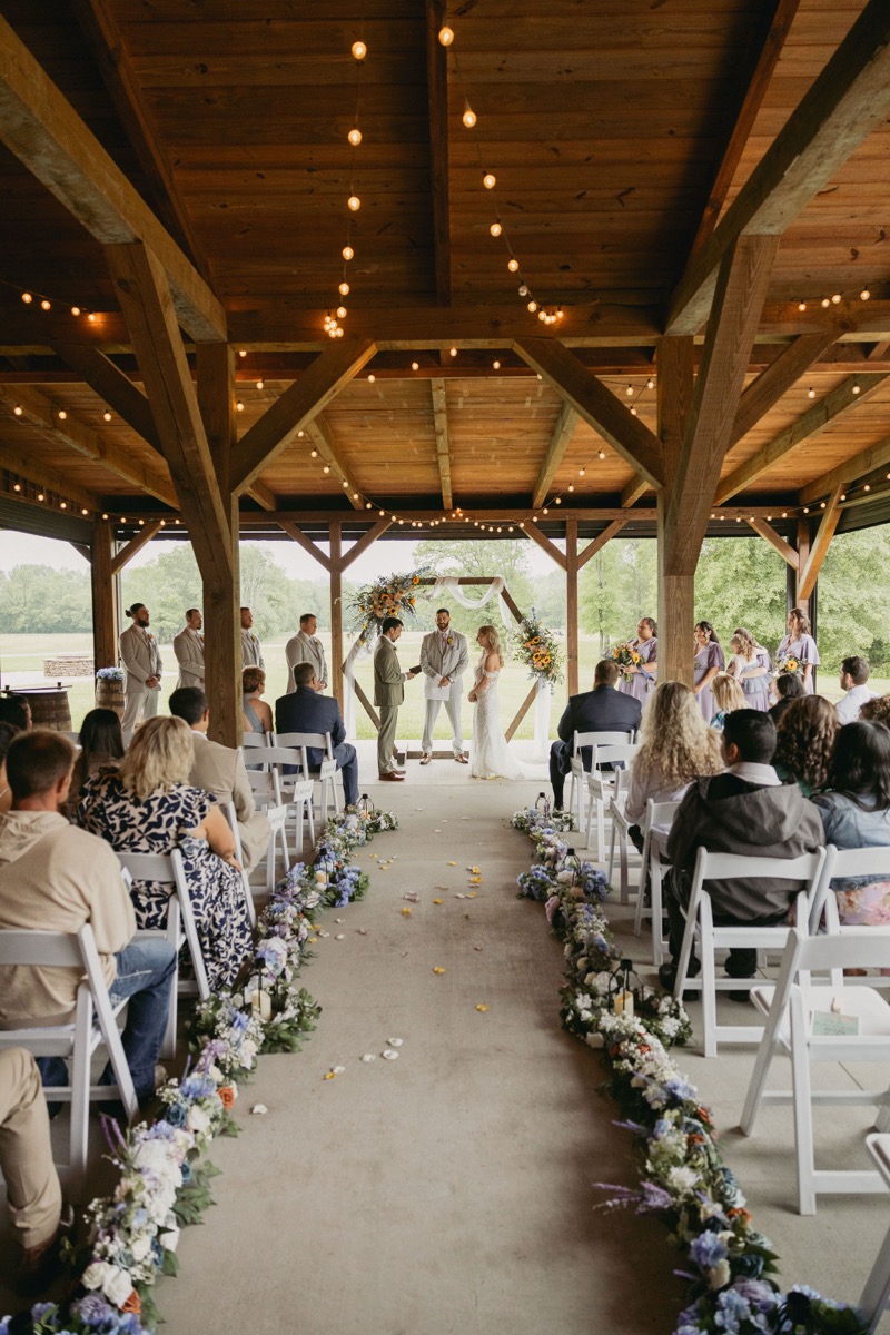 Brianna & Michael stand at the sunflower hexagonal arch down the petal-strewn aisle of Covey Creek's string-lit pavilion.