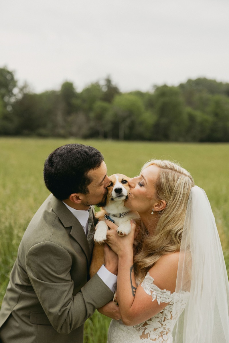 Brianna & Michael kiss their corgi between them in a green field under soft skies at Covey Creek.