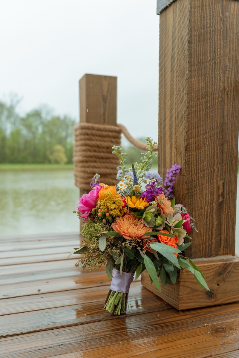 Caitlin & Daniel colorful bridal bouquet rests on the wet Pond dock by rope posts at Covey Creek