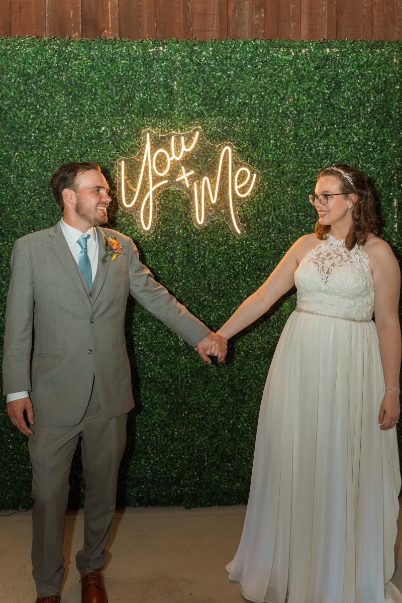 Caitlin & Daniel hold hands by a glowing You + Me neon on a boxwood wall indoors at Covey Creek