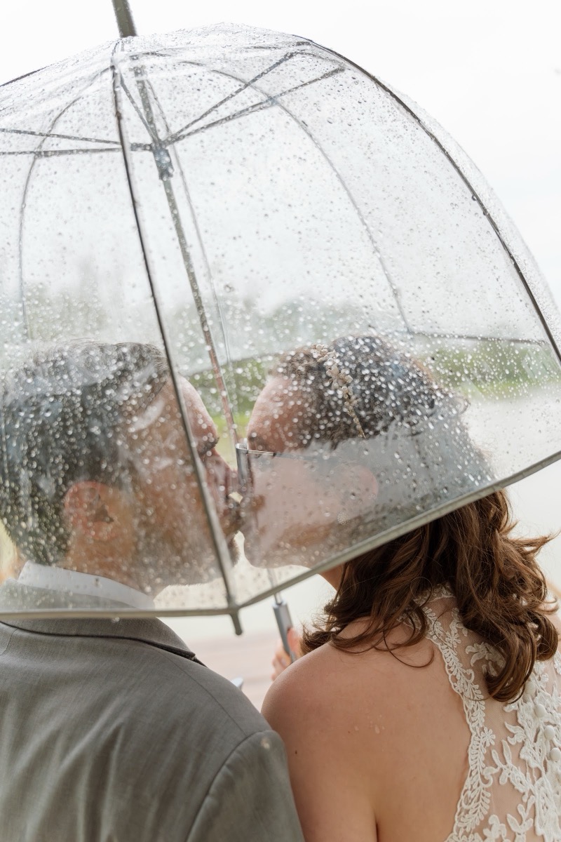 Caitlin & Daniel kiss beneath a rain-covered clear umbrella outdoors on their wedding day at Covey Creek