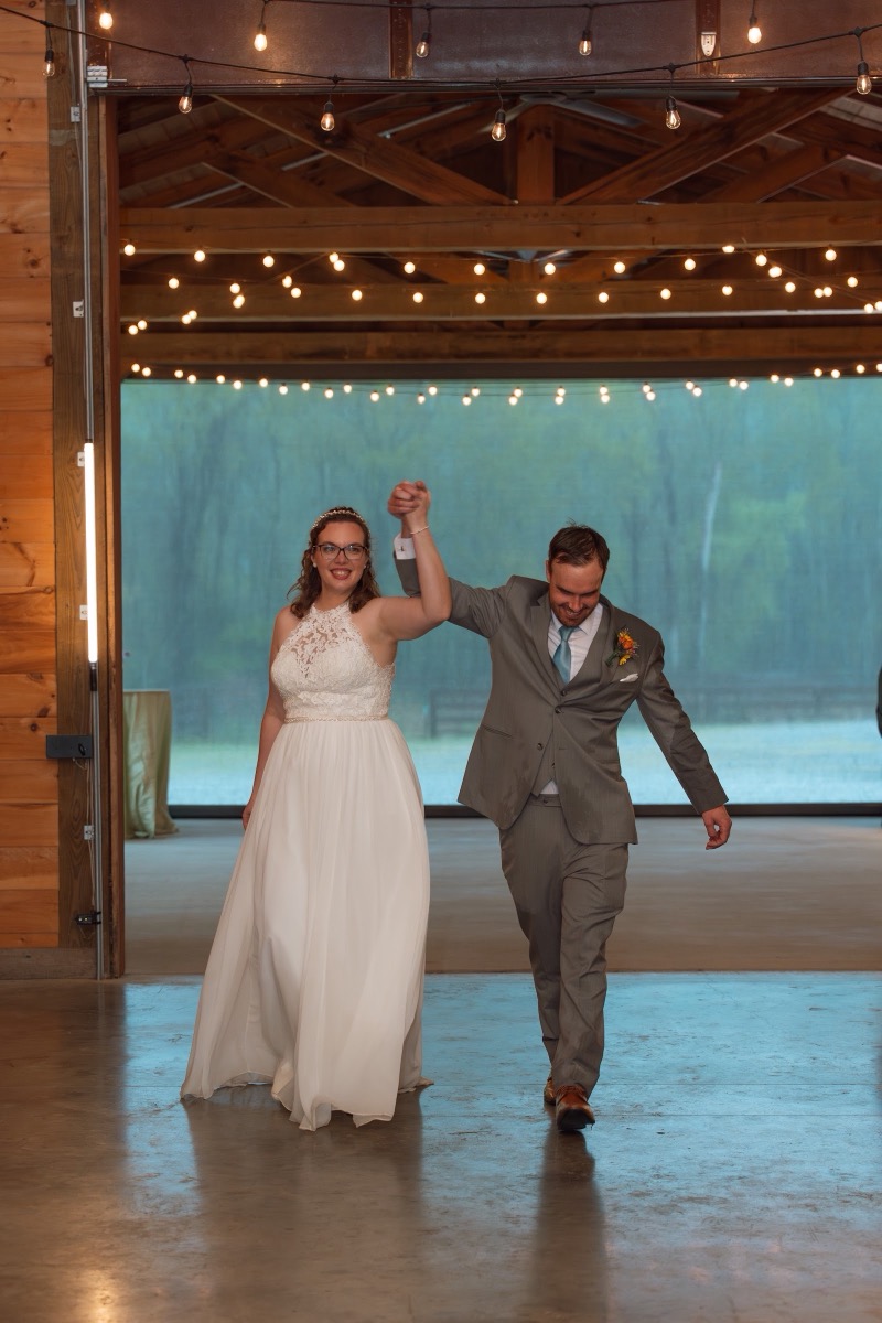 Caitlin & Daniel raise joined hands walking into the barn reception under string lights at Covey Creek