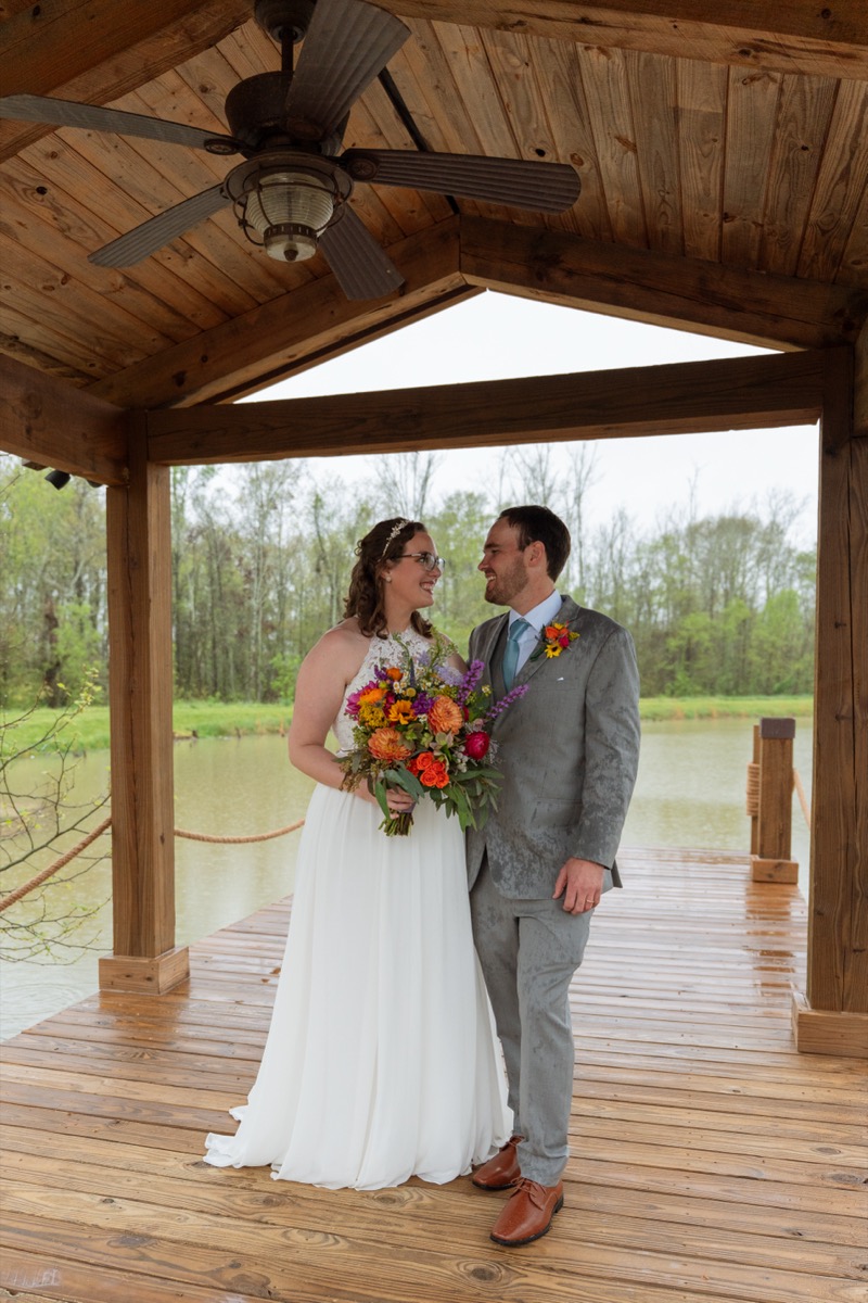 Caitlin & Daniel stand close on the wet Pond dock under a pavilion roof after rain at Covey Creek