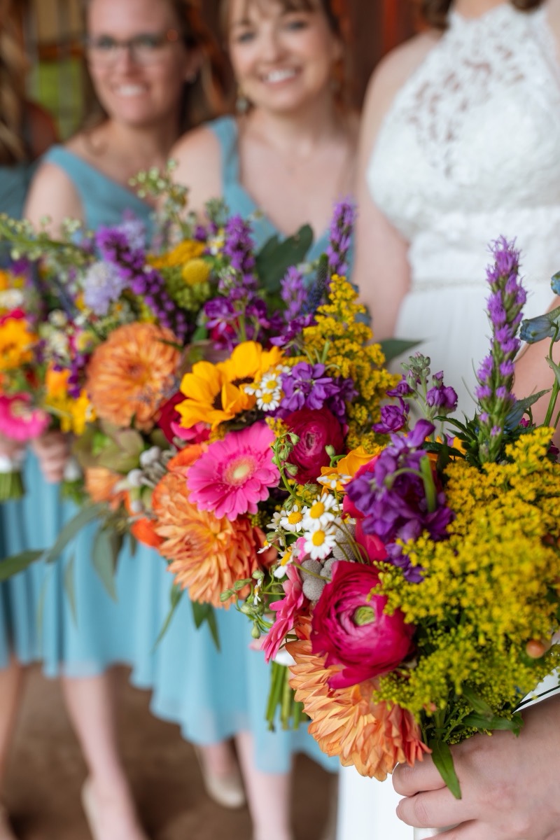 Caitlin & Daniel bridal party holds orange, pink, and yellow bouquets in dusty blue dresses at Covey Creek