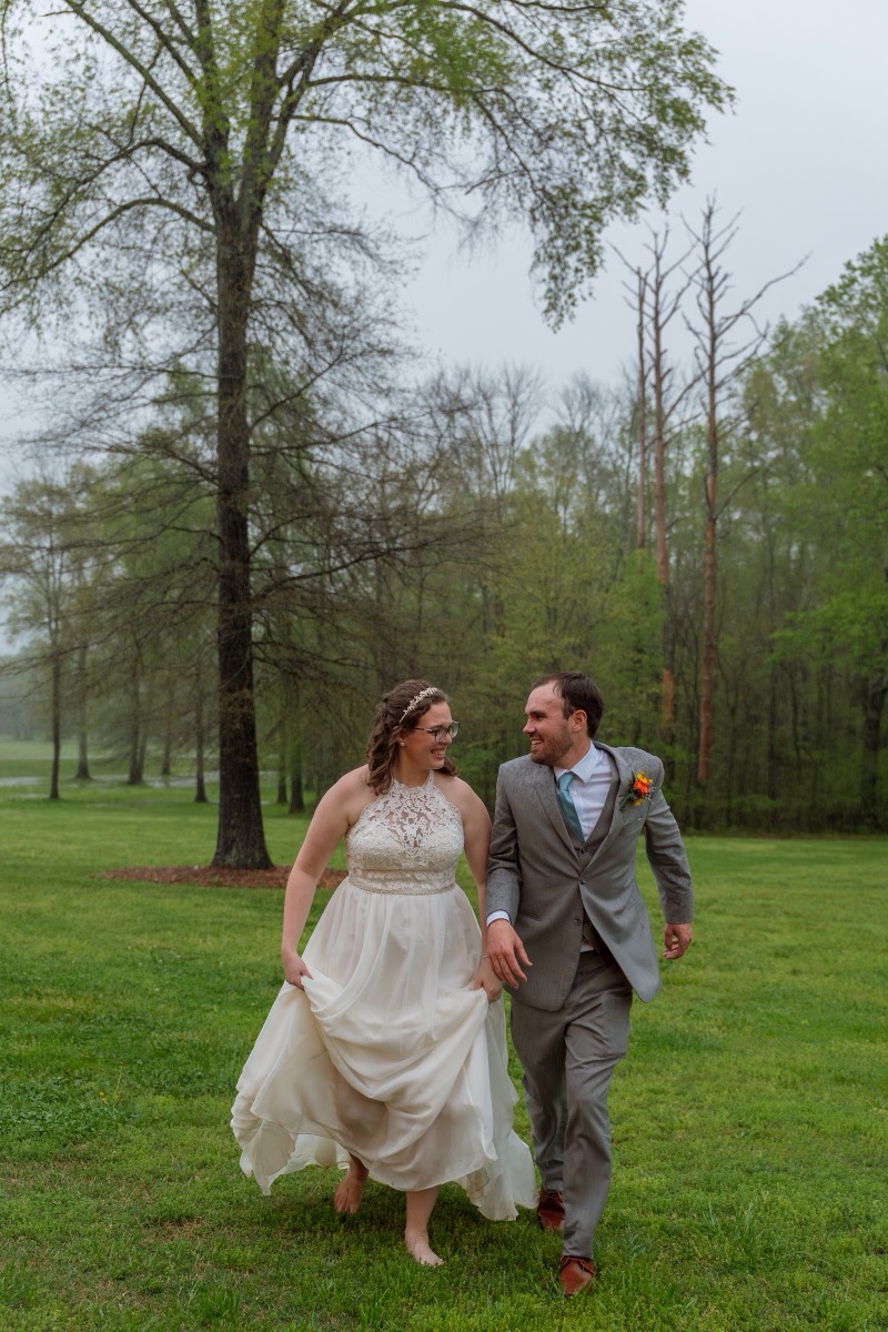 Caitlin & Daniel laugh barefoot running through a green spring field in gown and gray suit at Covey Creek
