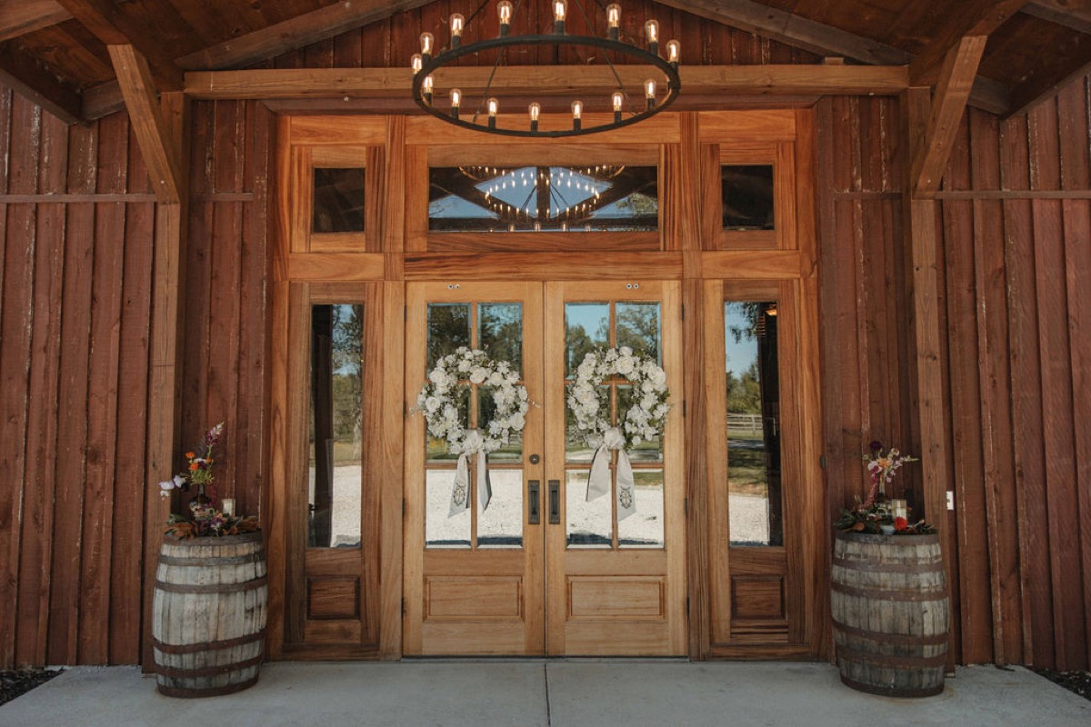 Floral wreaths hang on Covey Creek barn doors beside whiskey-barrel blooms for Claire & Jacob