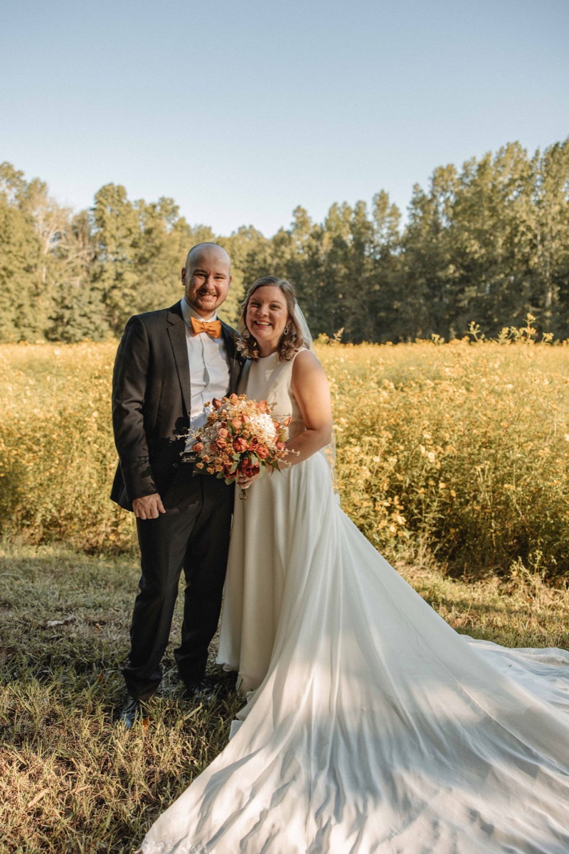 Claire & Jacob smile in a yellow wildflower meadow, orange bowtie and bridal bouquet at Covey Creek