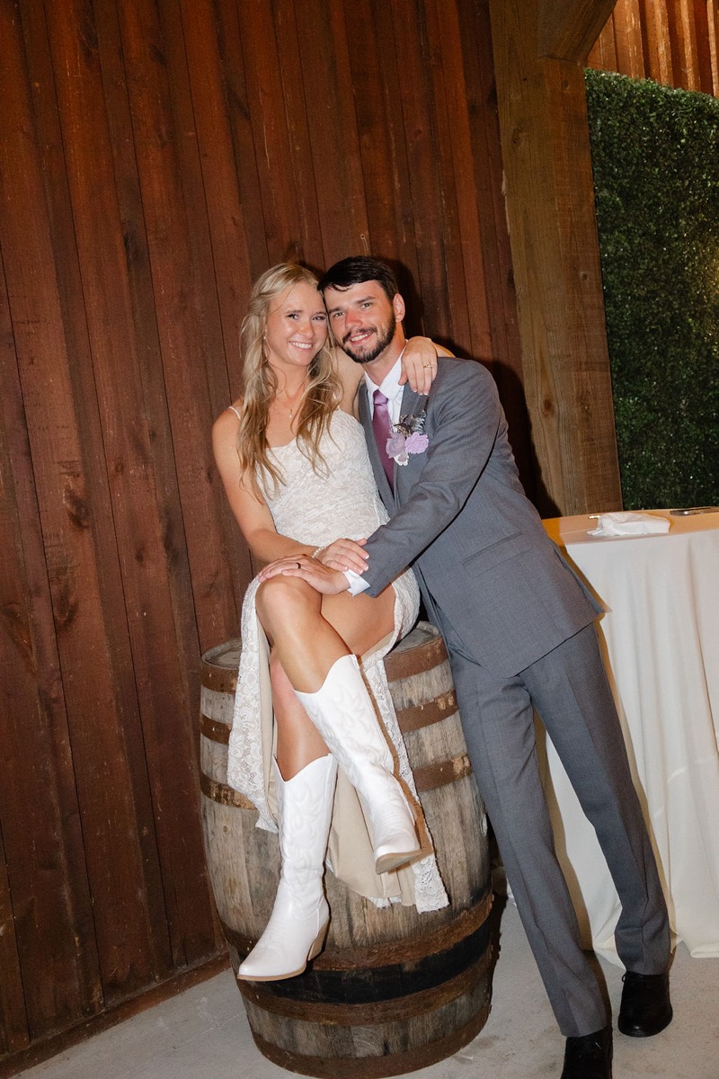 Courtney & Brett pose by a whiskey barrel and boxwood wall, her in white cowboy boots at Covey Creek