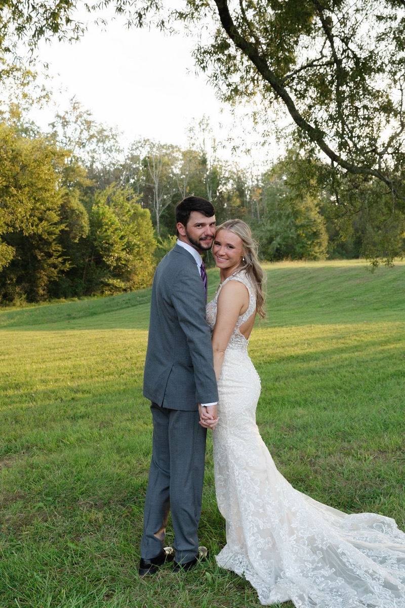 Courtney & Brett hold hands in a sunlit field, her lace train trailing on the grass at Covey Creek
