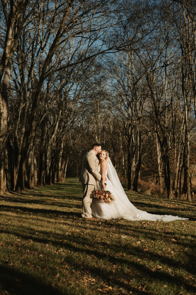 Danika & Troy stand in the Tunnel of Love tree path as he kisses her forehead at Covey Creek