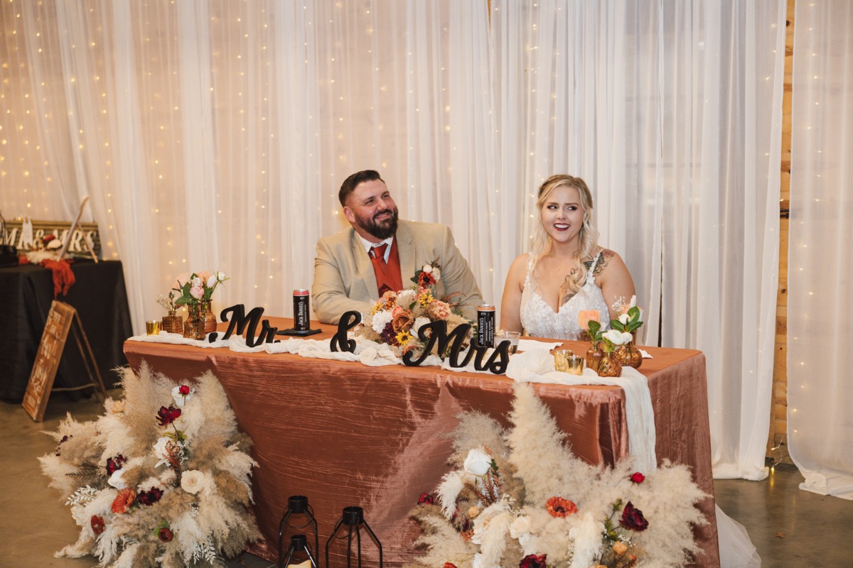 Danika & Troy smile at their rust Mr. & Mrs. sweetheart table with floor pampas at Covey Creek
