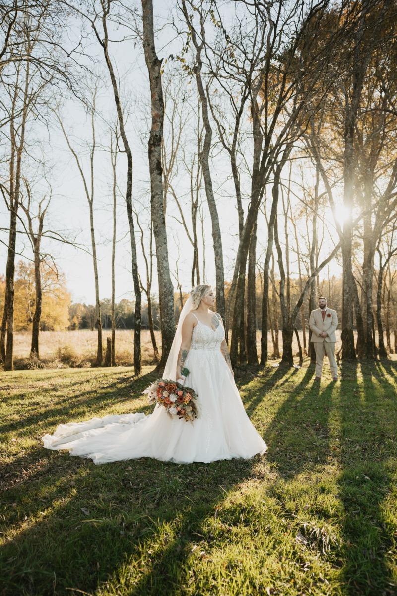 Danika & Troy pose at golden hour; she stands ahead in tulle as he waits among trees at Covey Creek