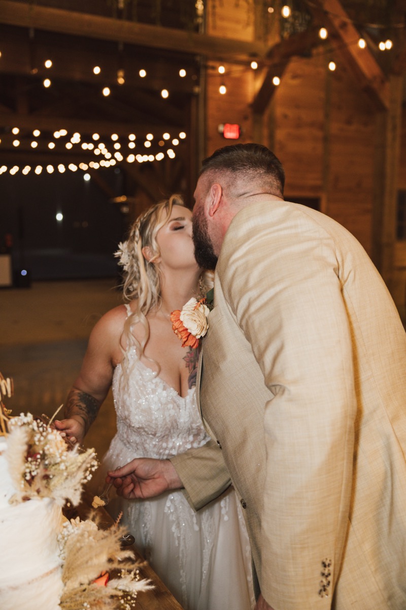 Danika & Troy kiss while cutting their autumn pampas-decorated cake in the barn at Covey Creek