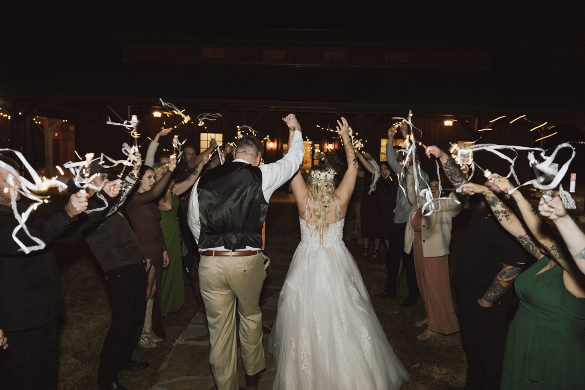 Danika & Troy raise joined hands walking through ribbon wands toward the barn at Covey Creek
