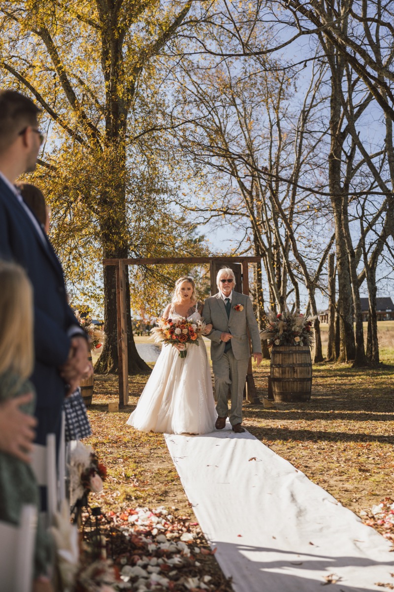Danika & Troy's processional: she walks the white aisle with her escort under autumn trees at Covey Creek