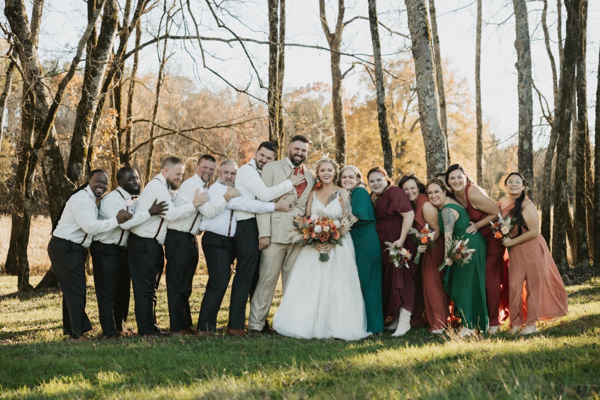 Danika & Troy laugh with their wedding party huddled close in a sunlit field at Covey Creek