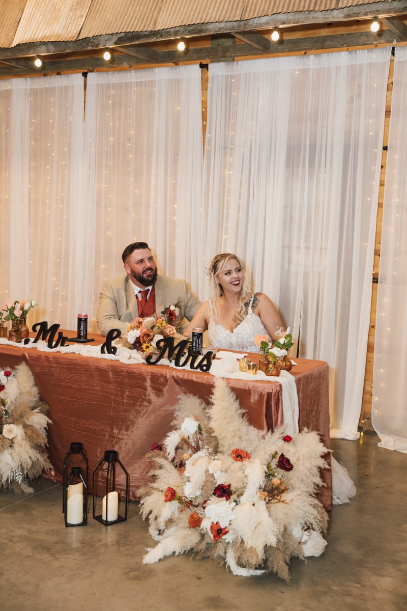 Danika & Troy sit smiling at their rust sweetheart table under globe lights at Covey Creek