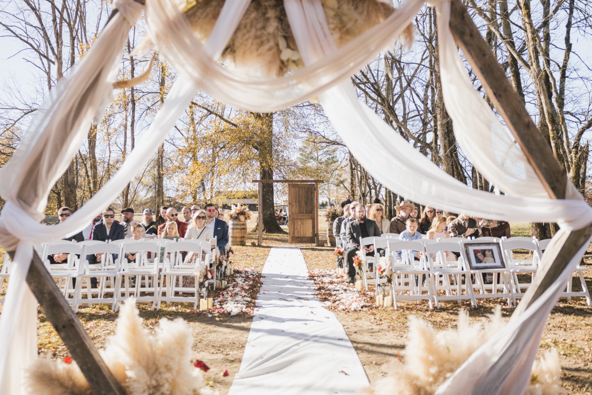 Guests fill white chairs beyond Danika & Troy's fabric-draped triangular arch aisle at Covey Creek