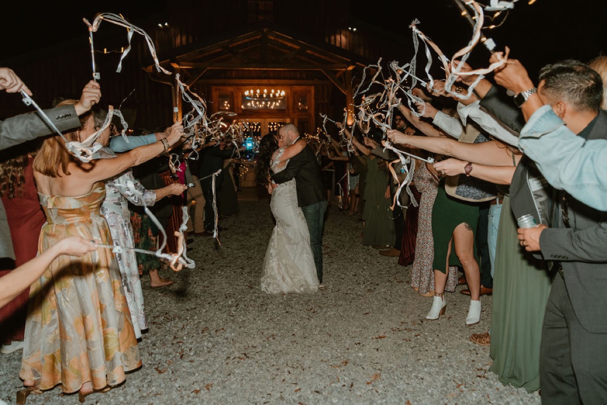 Elizabeth & Ashton kiss on the gravel at Covey Creek as guests form a lit ribbon-wand tunnel by the barn