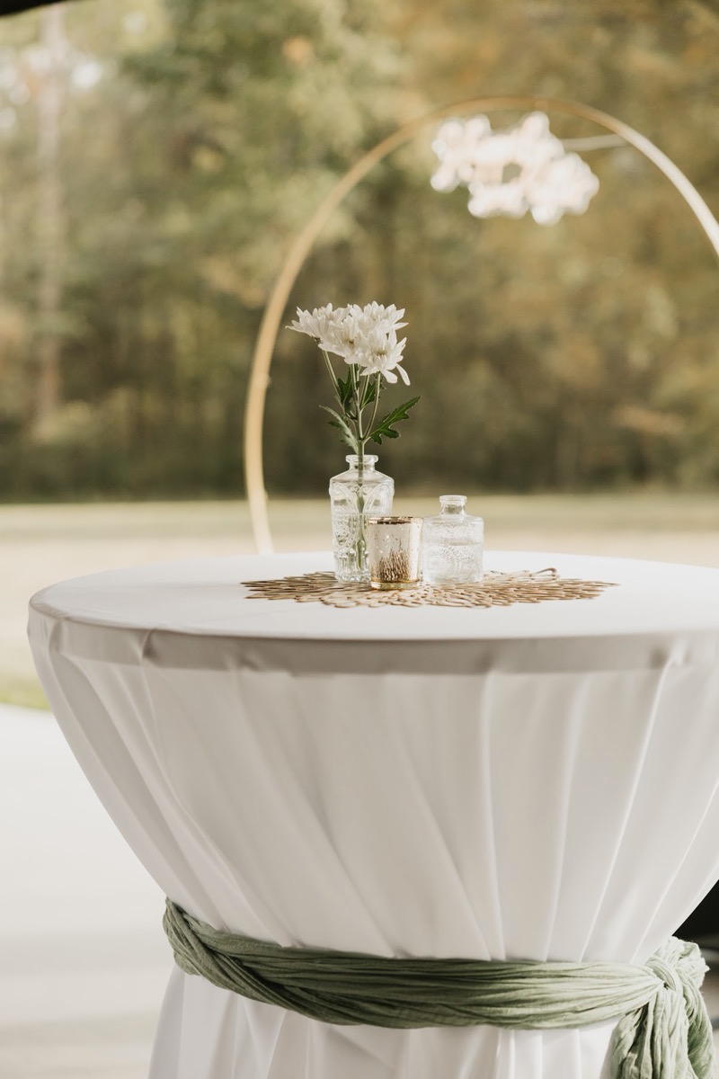 A cinched cocktail table with white blooms and a gold moon arch sits on the Patio for Emily & Bryan's reception at Covey Creek.