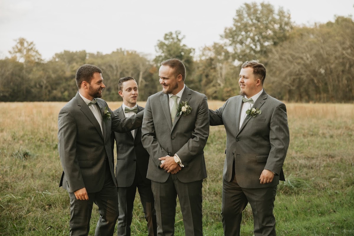 Bryan laughs with groomsmen in matching grey suits and sage ties in a golden field during Emily & Bryan's day at Covey Creek.