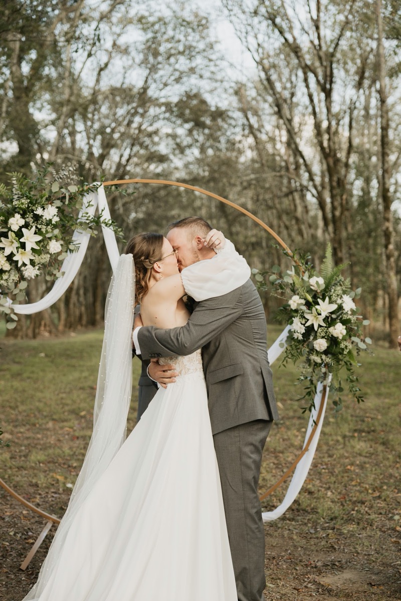 Emily & Bryan share their first kiss before a circular floral arch under the Tunnel of Love trees at Covey Creek.