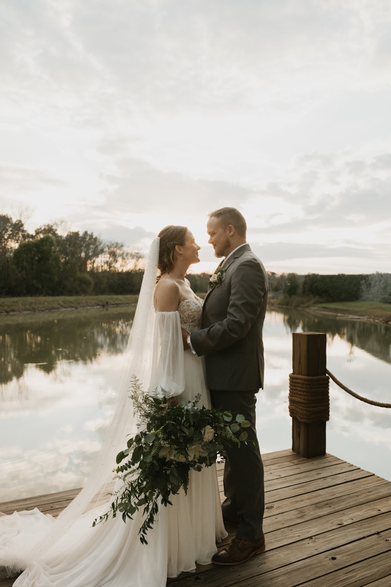 Emily & Bryan stand face-to-face on a wooden dock by the Pond, her cascading bouquet between them at Covey Creek.