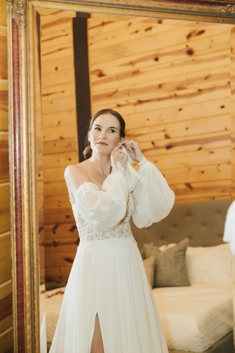 Emily adjusts an earring in a gold-framed mirror in the bridal suite, lace sleeves reflected at Covey Creek for Emily & Bryan.