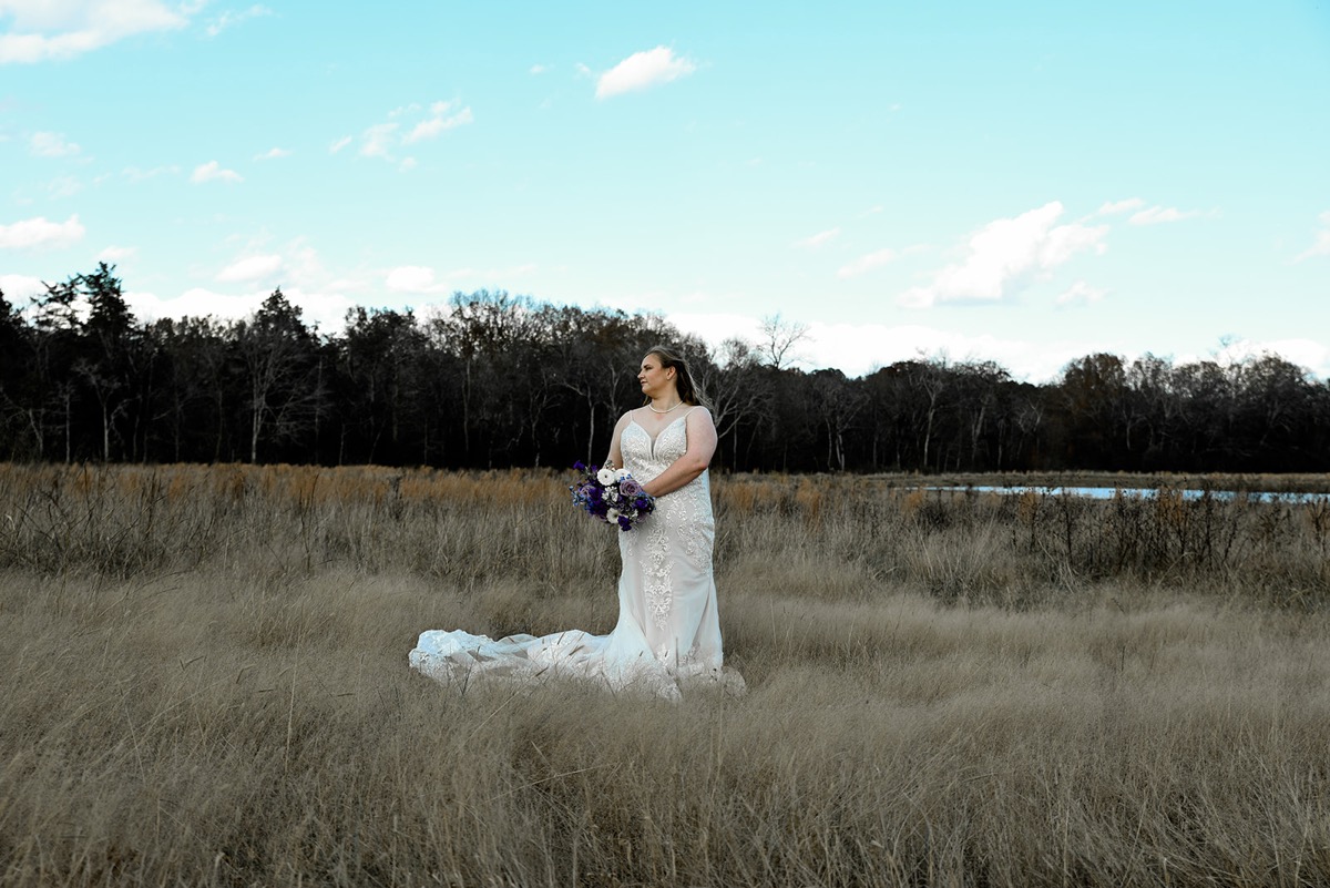 Emily stands alone in a tall grassy field in her lace gown, purple bouquet in hand, for Emily & Jared at Covey Creek.