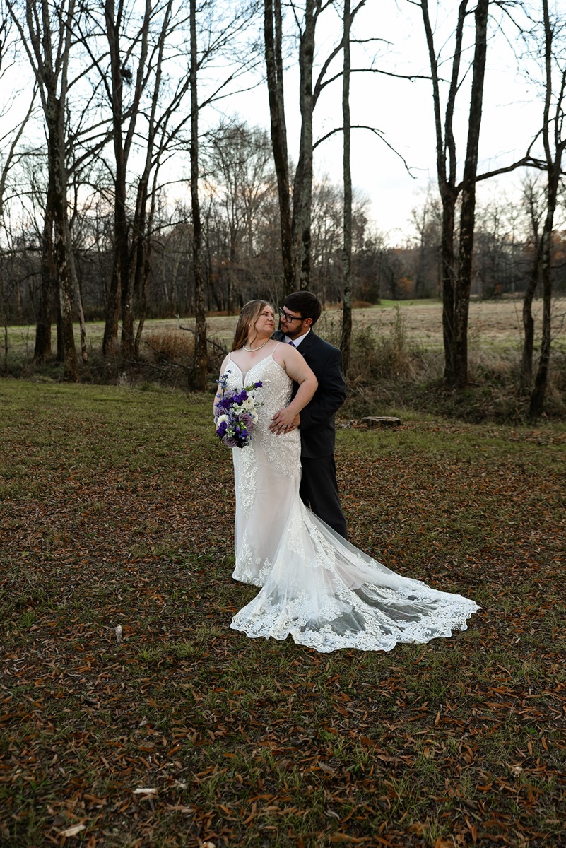 Emily & Jared face each other in a leaf-strewn clearing, her purple bouquet and lace train spread at Covey Creek.
