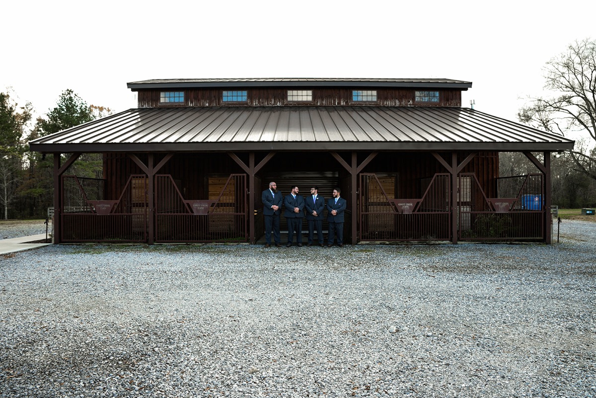 Jared and three groomsmen in navy suits stand under the barn porch for Emily & Jared's wedding at Covey Creek.