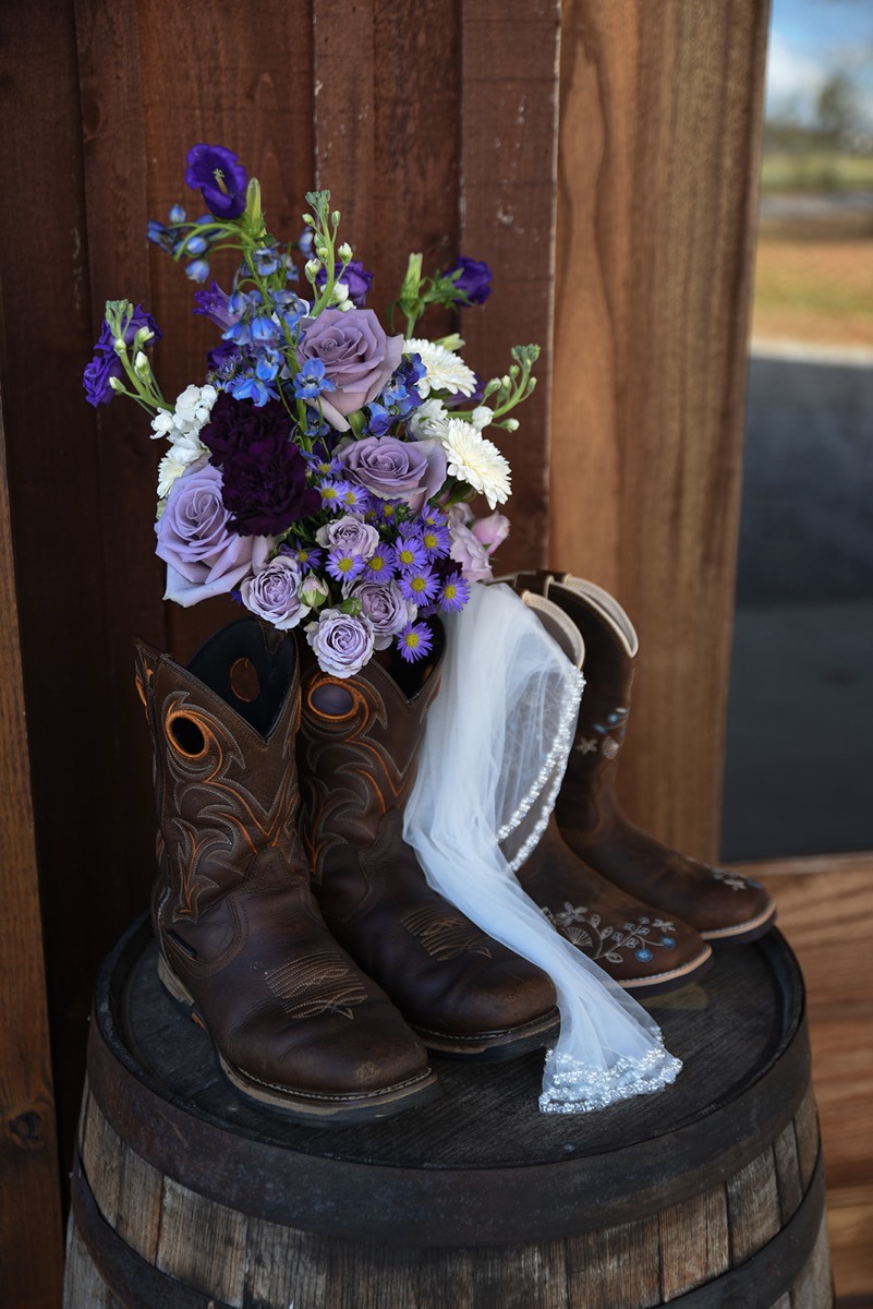 His-and-hers cowboy boots, a purple bouquet, and a beaded veil rest on a barrel for Emily & Jared at Covey Creek.