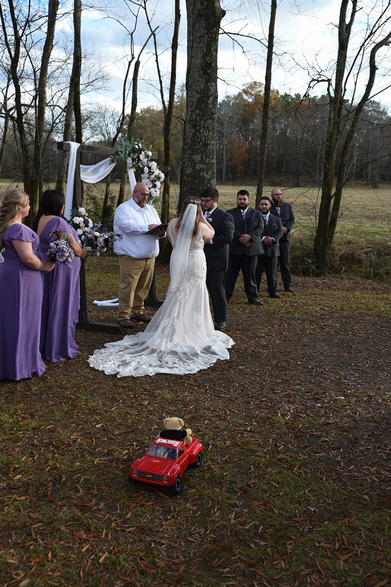 From behind, Emily & Jared stand at a flowered arbor while a red toy truck rests in the aisle at Covey Creek.