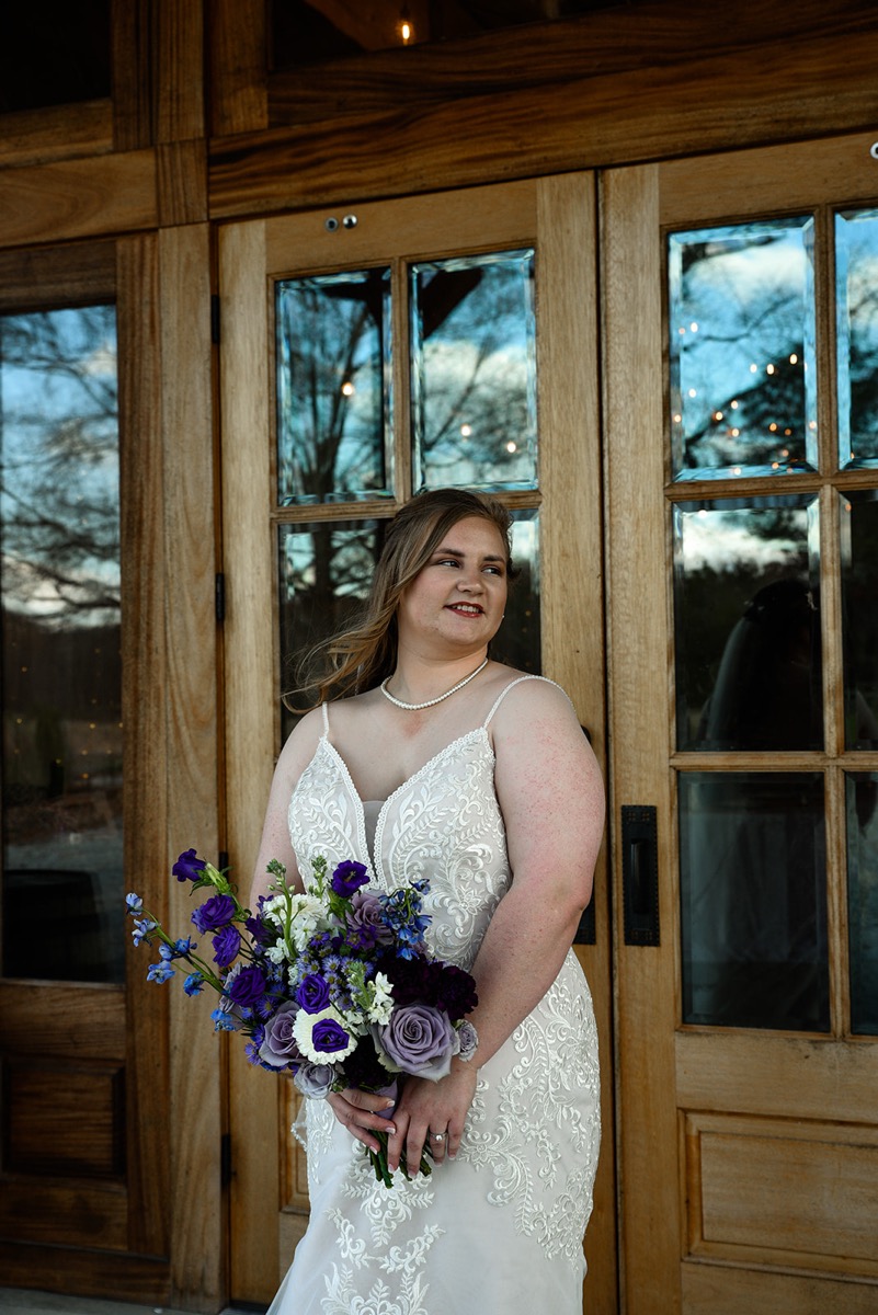Emily smiles with her purple bouquet before glass-paneled wooden doors for Emily & Jared's day at Covey Creek.