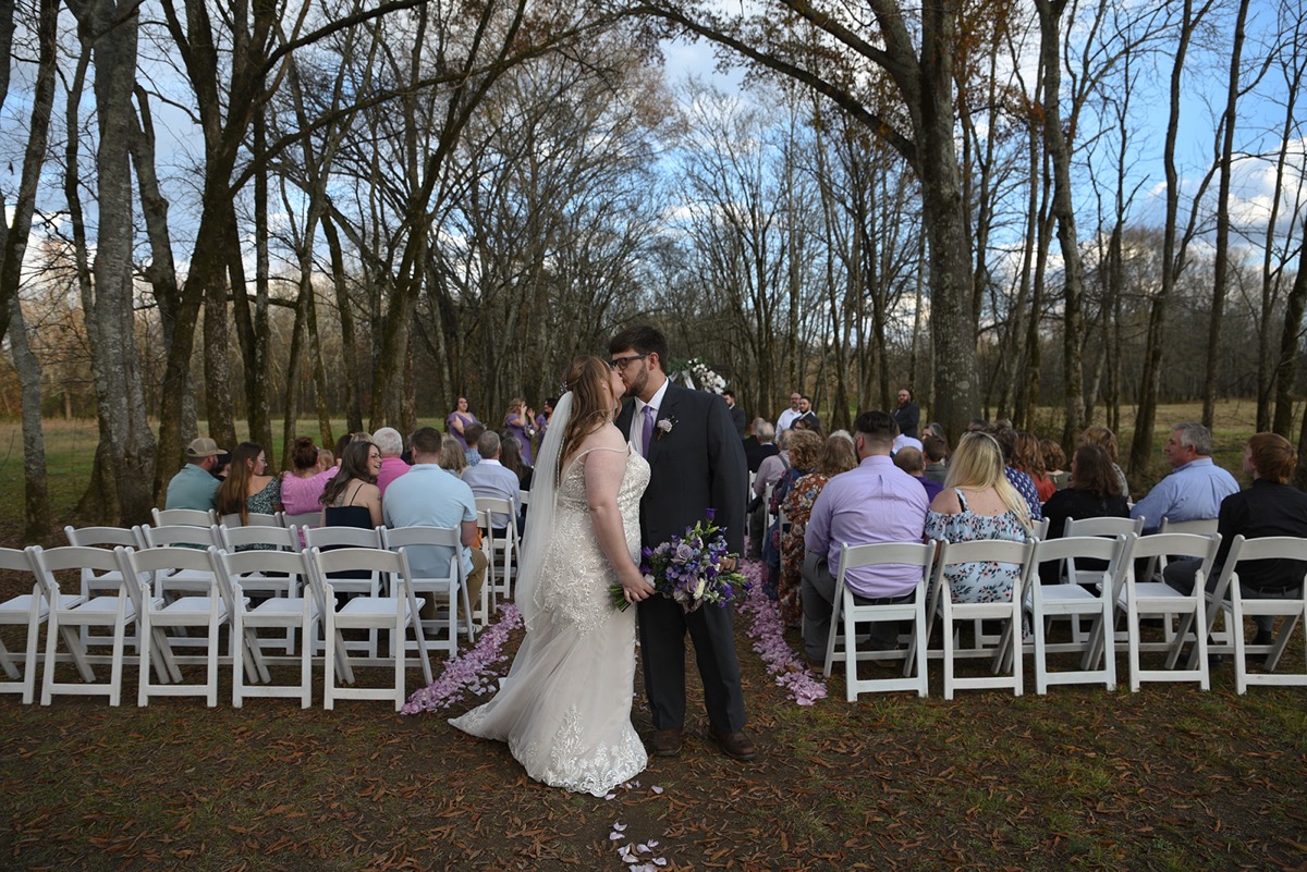 Emily & Jared share a kiss in the wooded aisle after vows as seated guests look on at Covey Creek.