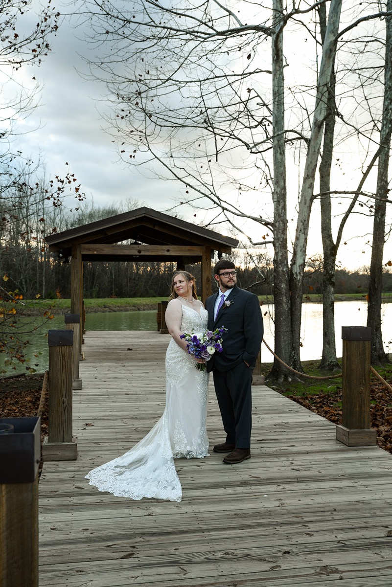Emily & Jared stand on the Pond dock by a wooden pavilion, her lace train on the boards at Covey Creek.