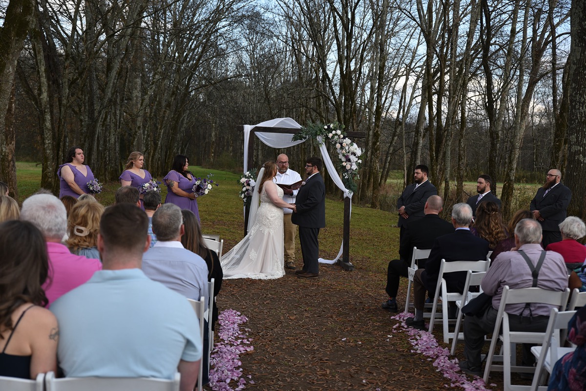 Emily & Jared exchange vows under a flowered wooden arbor as guests watch their wooded ceremony at Covey Creek.