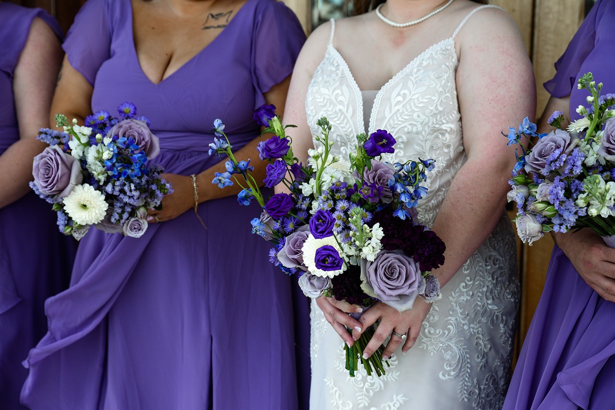 Emily's purple and white bouquet fills the frame beside bridesmaids in lavender for Emily & Jared at Covey Creek.