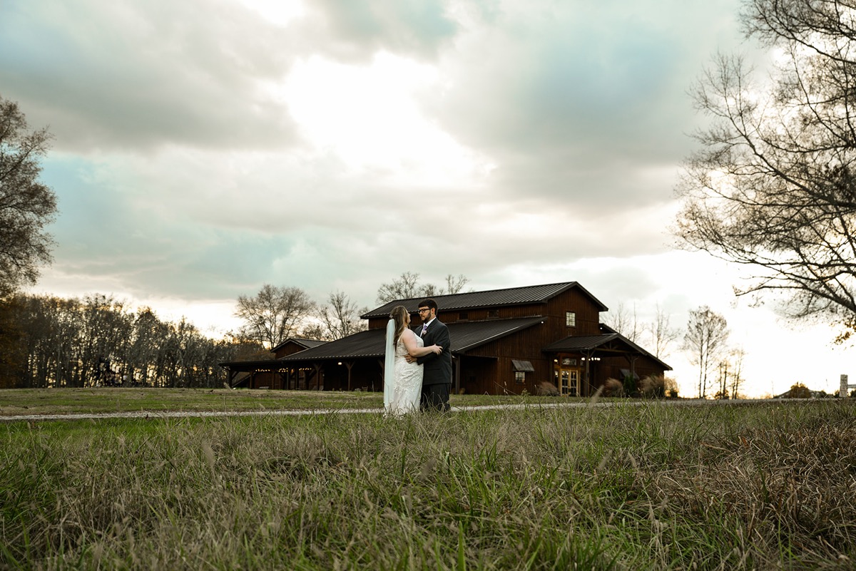 Emily & Jared embrace in tall grass before the rustic barn under a cloudy sky at Covey Creek.