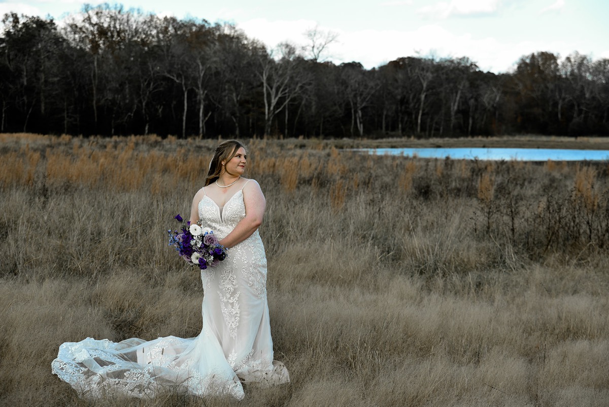 Emily glances over her shoulder in a lace gown near the Pond with a purple bouquet for Emily & Jared at Covey Creek.