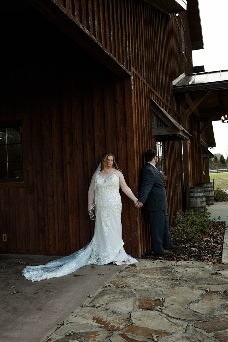 Emily & Jared share a first touch around a barn corner, her lace train fanned on stone at Covey Creek.