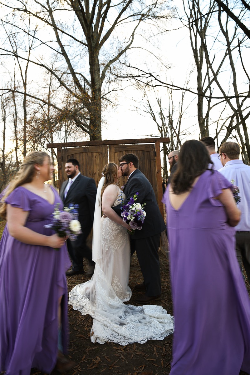 Emily & Jared kiss by a wooden doorway as bridesmaids in purple pass through fallen leaves at Covey Creek.