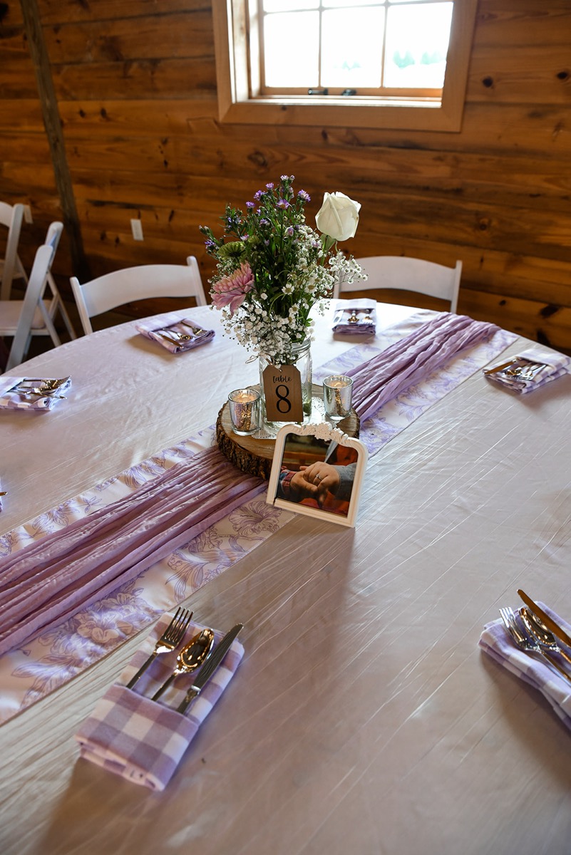 Table 8 at Emily & Jared's reception holds purple florals, gingham napkins, and a framed photo indoors at Covey Creek.