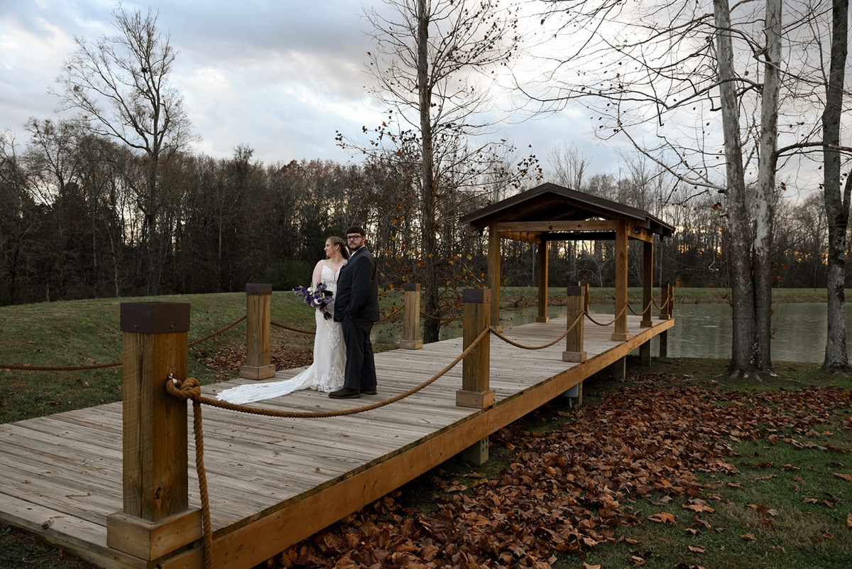 Emily & Jared pose on the Pond dock, her lace train on the boards and a pavilion behind them at Covey Creek.