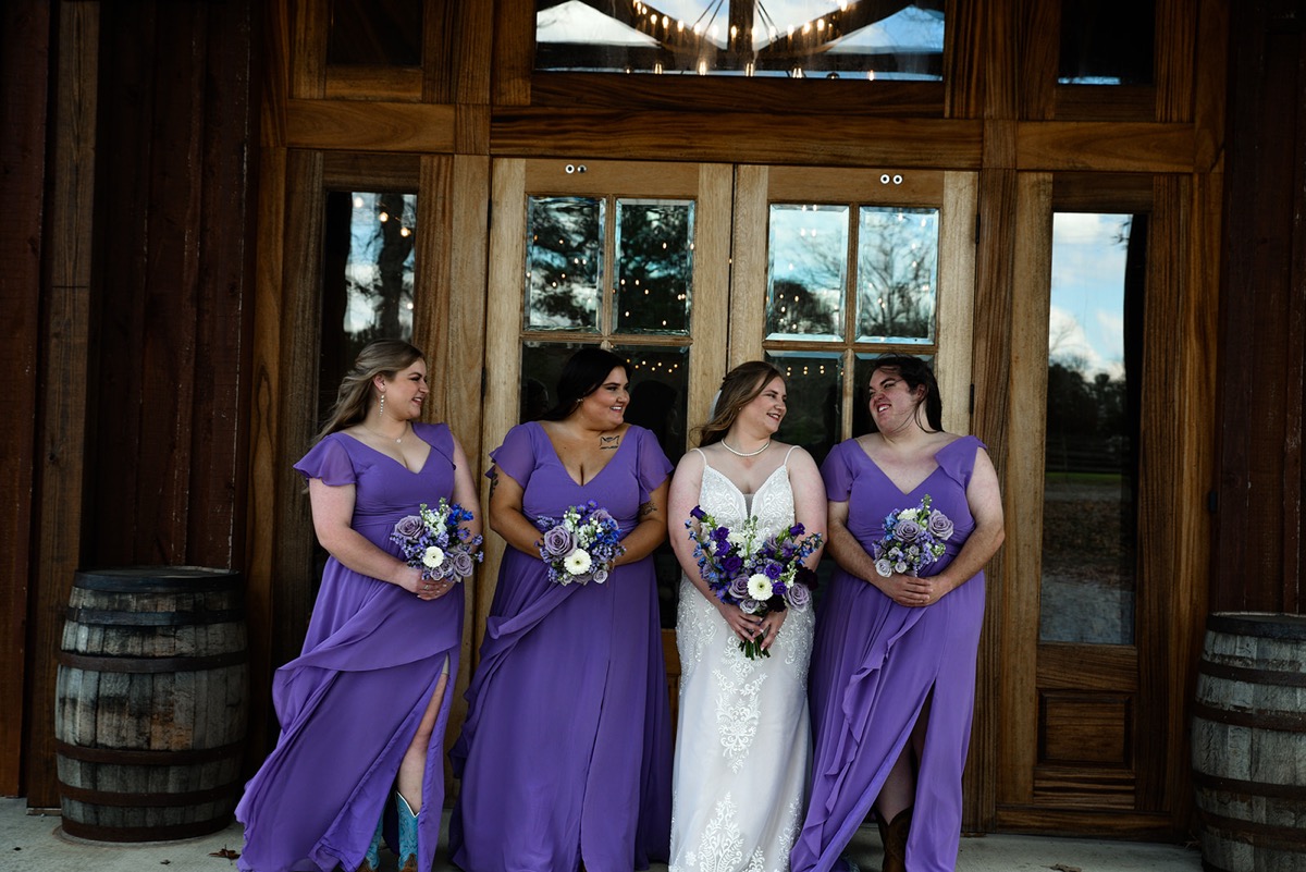 Emily and three bridesmaids in lavender gowns laugh by wooden doors for Emily & Jared's wedding at Covey Creek.