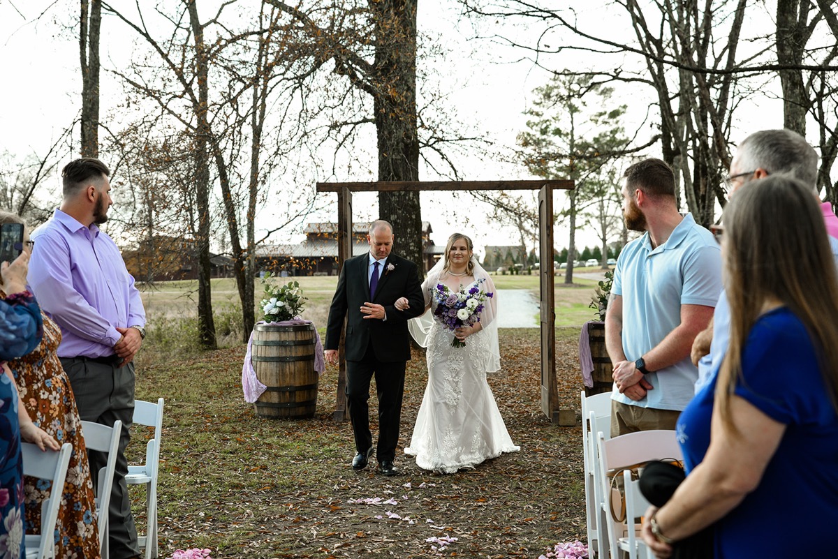 Emily walks the petal-strewn aisle on her father's arm toward their wooded ceremony for Emily & Jared at Covey Creek.