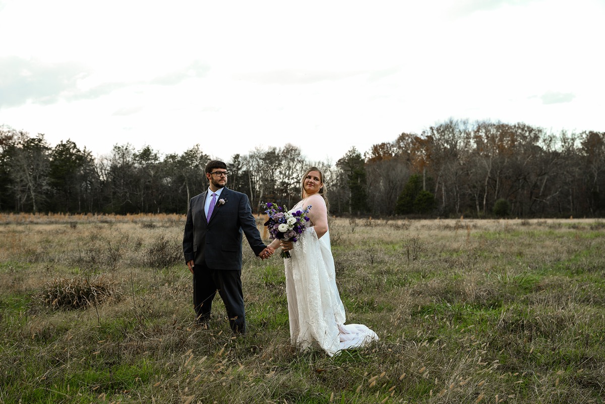 Emily & Jared hold hands in a tall grassy field, she glancing back with her purple bouquet at Covey Creek.