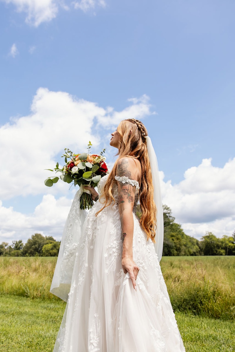 Erica & Randall’s bride looks skyward in lace with a red bouquet on the grassy field at Covey Creek