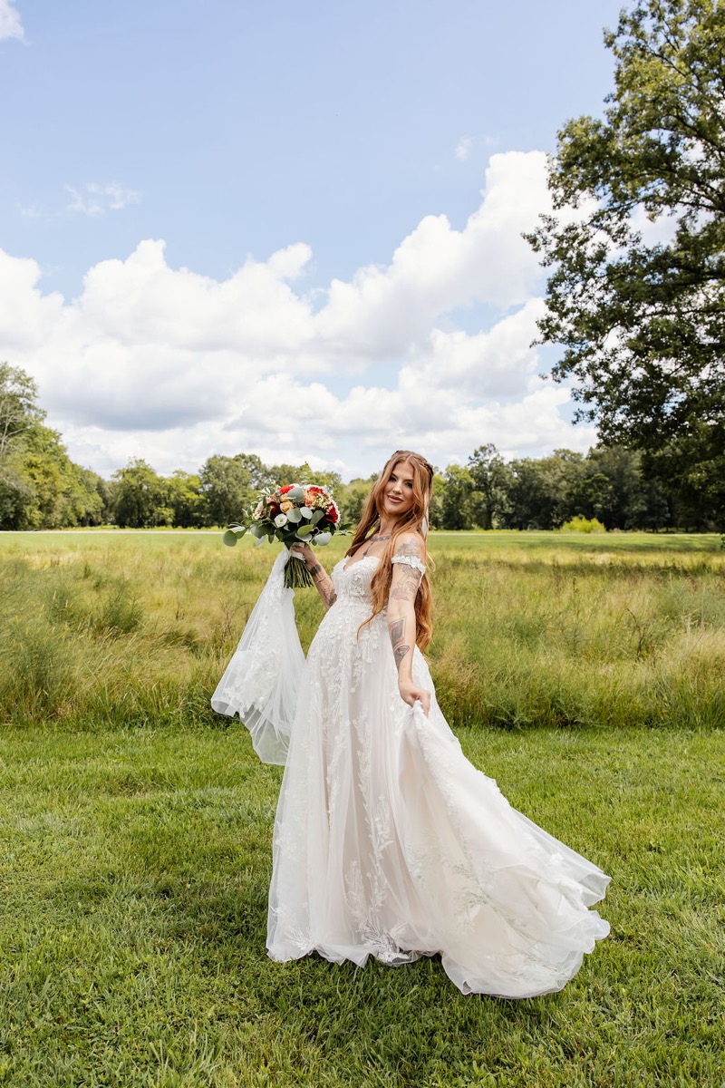 Erica & Randall’s bride stands in lace with a red-orange bouquet against the Covey Creek field and blue sky